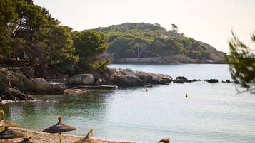 A beach with thatched umbrellas, calm water, rocky coastline, and a hill with trees.