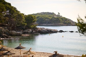 A beach with thatched umbrellas, calm water, rocky coastline, and a hill with trees. - Aluasoul Hotel Carolina (Capdepera)