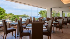 A restaurant with tables and chairs arranged for dining, large windows offering a view of the sea and greenery. - Aluasoul Hotel Carolina (Capdepera)