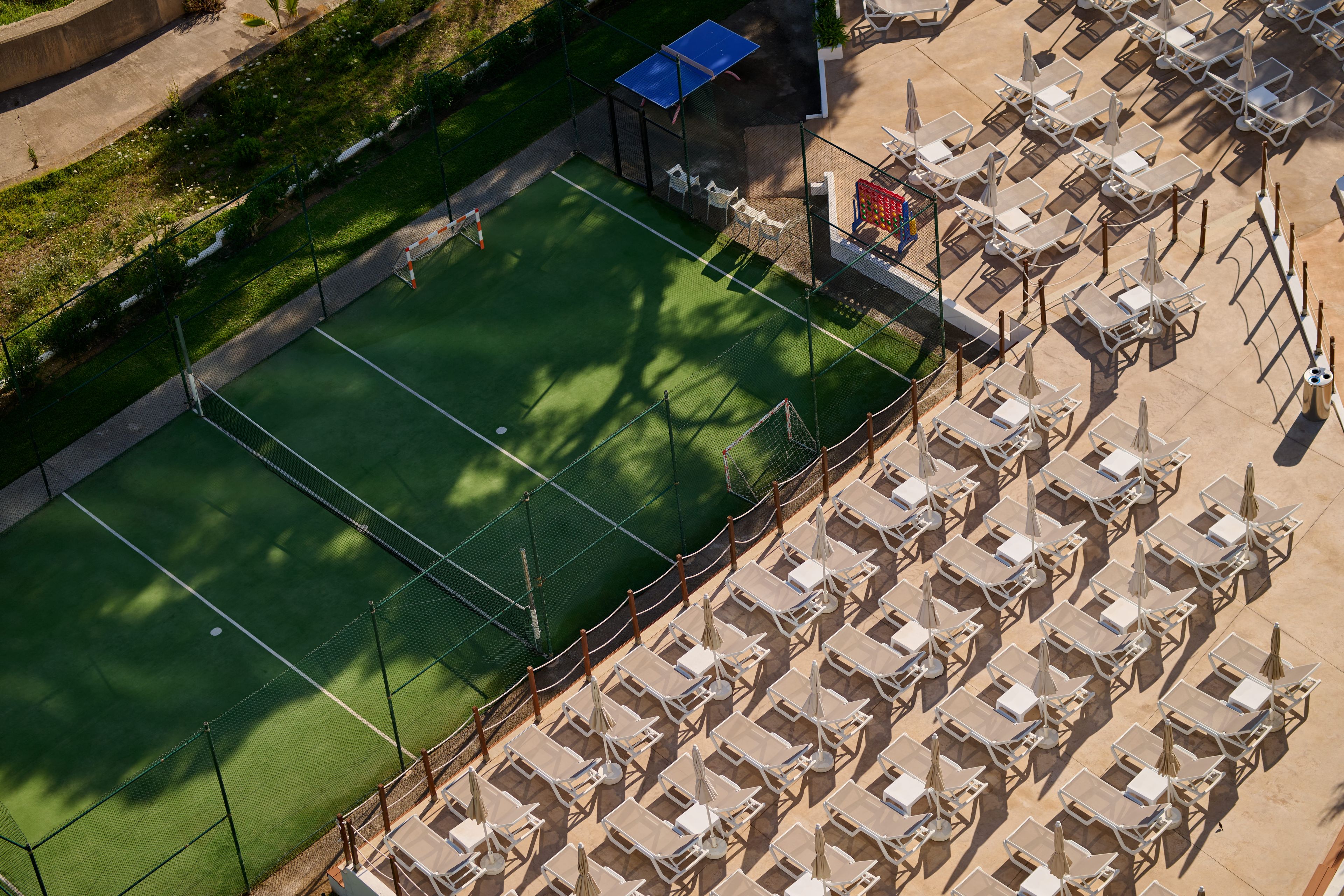 Vista aerea di un campo da tennis circondato da lettini e ombrelloni, con un piccolo edificio adiacente al campo.