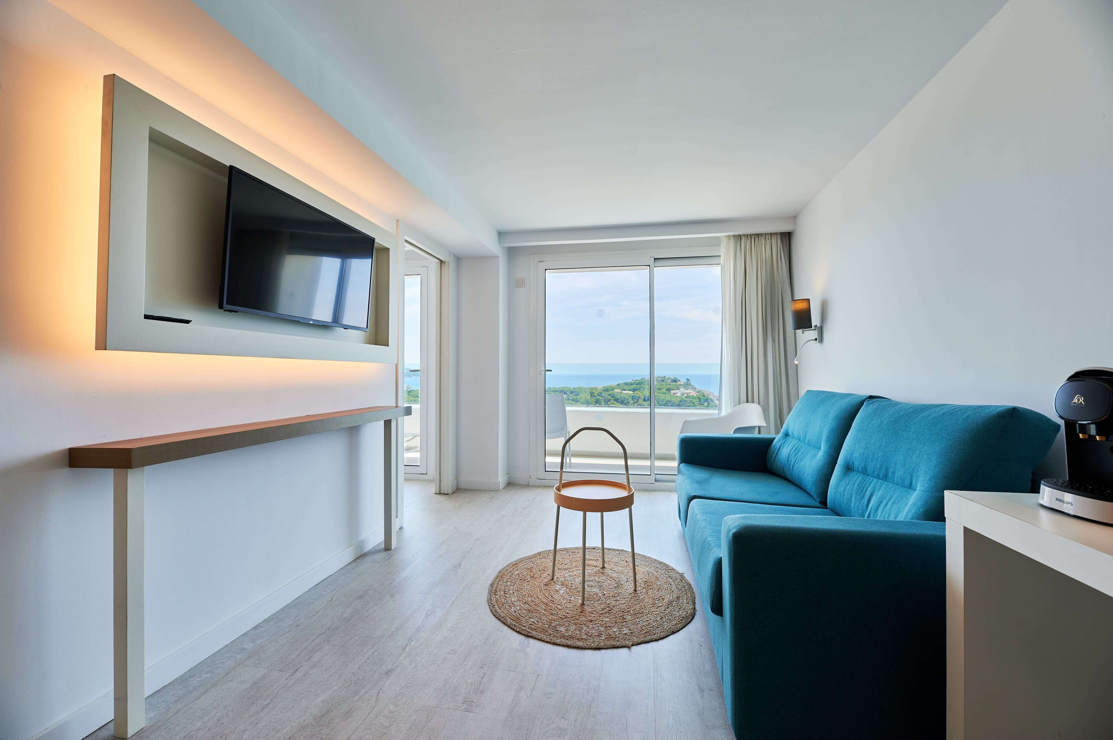 A modern hotel room with a flat-screen TV mounted on the wall, a teal sofa, a small round table, and a view of the sea through large windows.
