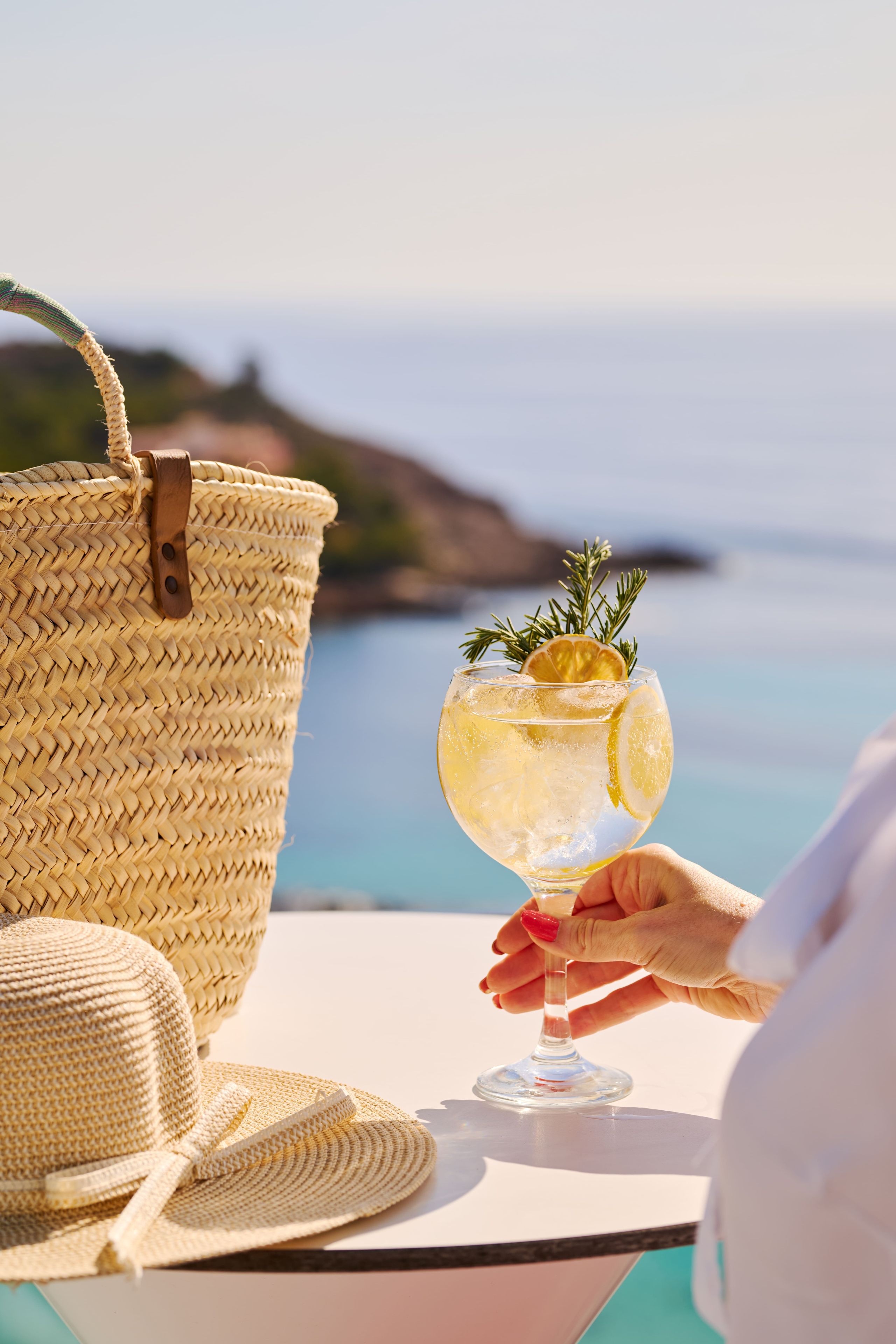 Un bolso de paja, un sombrero de paja y un vaso de bebida sobre una mesa con vistas al mar.