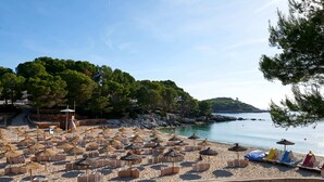 A beach with rows of sun loungers and umbrellas, a lifeguard tower, and a rocky coastline with trees. - Aluasoul Hotel Carolina (Capdepera)