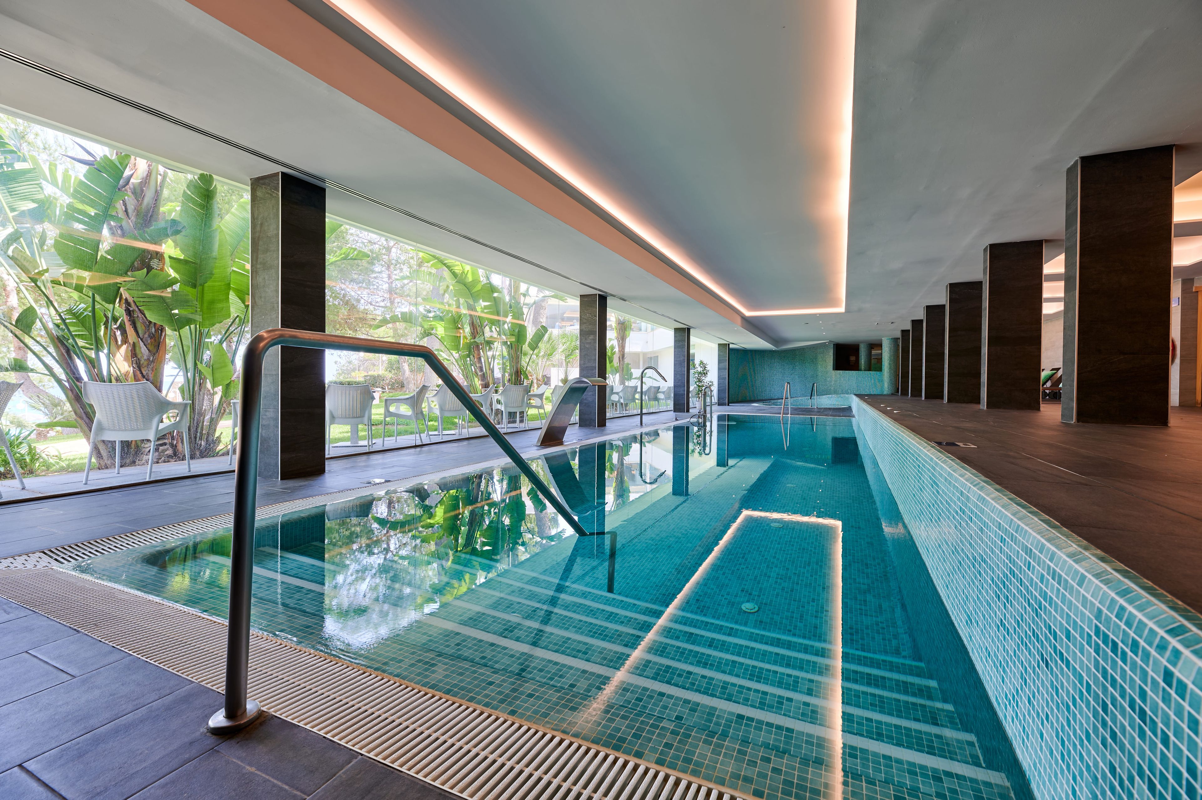 Ein moderner, überdachter Swimmingpool mit großen Glaswänden, Fliesenboden und Blick auf Grünflächen.