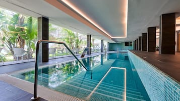 Una piscina interna moderna con pareti in vetro trasparente, pavimento piastrellato e vista sul verde circostante.
