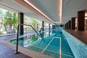 A modern indoor swimming pool with clear glass walls, a tiled floor, and a view of greenery outside. - Aluasoul Hotel Carolina (Capdepera)