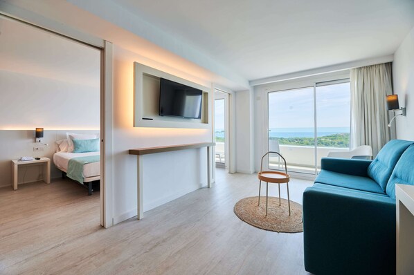 A modern hotel room with a bed, a TV mounted on the wall, a sofa, and a large window offering a view of the sea. - Aluasoul Hotel Carolina (Capdepera)