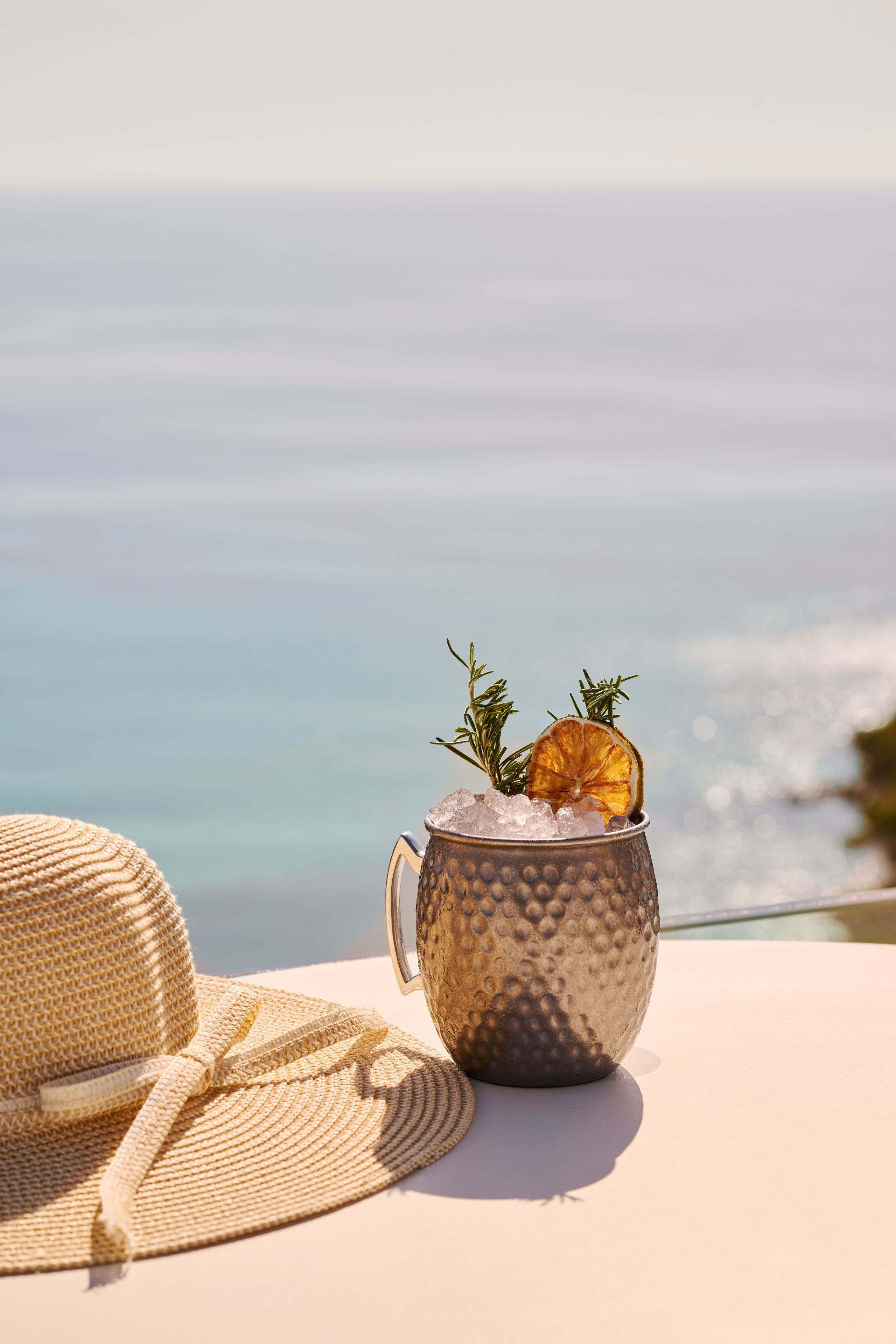 A woven straw hat and a textured mug with ice and a dried orange slice on a table with a view of the sea.