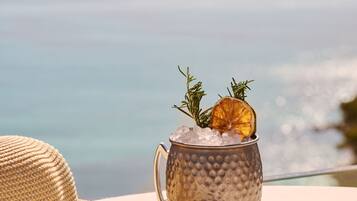 A woven straw hat and a textured mug with ice and a dried orange slice on a table with a view of the sea.