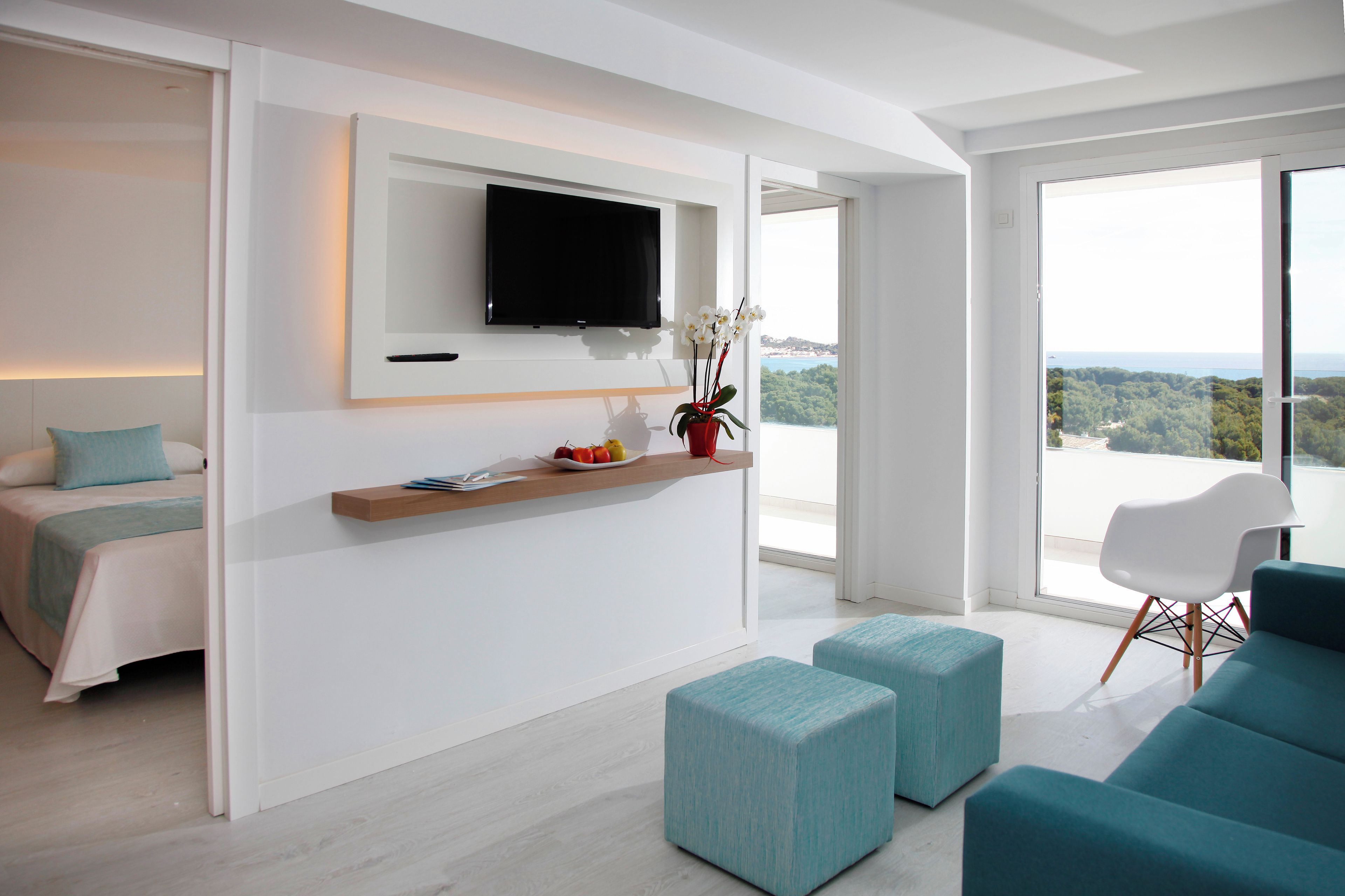 A modern hotel room with a bed, a TV mounted on the wall, a small table with a plant, and a view of the outdoors through large windows.