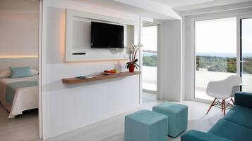 A modern hotel room with a bed, a TV mounted on the wall, a small table with a plant, and a view of the outdoors through large windows.