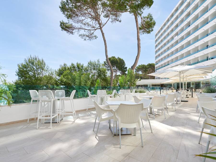 Un patio exterior con mobiliario de mimbre blanco, barandilla de vidrio y vista a un edificio moderno.