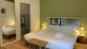 Une chambre à coucher avec un lit, des lampes de chevet, un petit banc en bois, un miroir et une fenêtre donnant sur un paysage agréable.