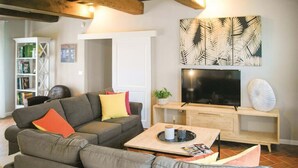 Ein modernes Wohnzimmer mit einem grauen Ecksofa, einem Couchtisch mit Zeitschriften, einem Flachbildfernseher auf einem Holzständer und einem Bücherregal im Hintergrund.