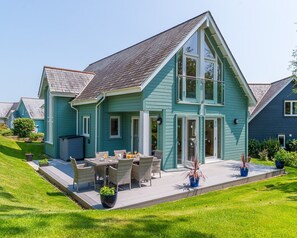 Ein zweistöckiges Haus mit geneigtem Dach, einer überdachten Terrasse mit Gartenmöbeln und einem gepflegten Rasen.
