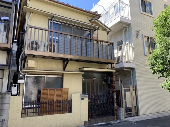 Un edificio residencial de dos pisos con balcón, entrada con portón y unidad de aire acondicionado visible.