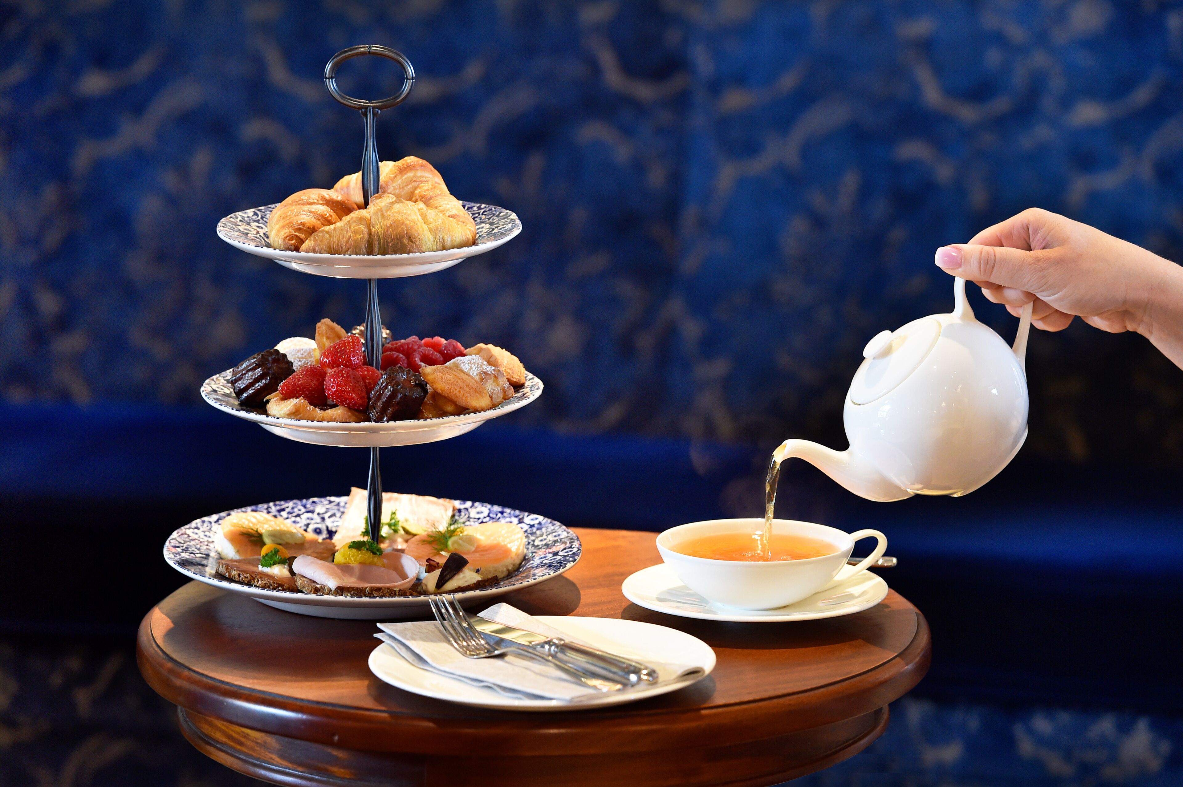 A tiered dessert stand with pastries and fruits, a teapot pouring tea, and dining utensils on a table.