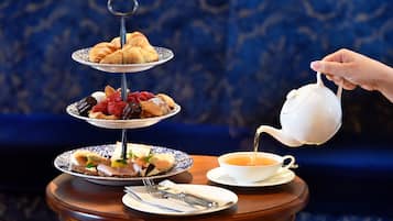 A tiered dessert stand with pastries and fruits, a teapot pouring tea, and dining utensils on a table.