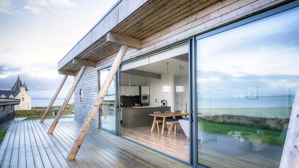 Ein modernes Haus mit einer Holzterrasse, einem Essbereich mit Stühlen und einer Küche.