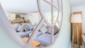 A modern living room with a grey sofa and a wooden coffee table. A large mirror reflects the room, and a lamp is on a side table.