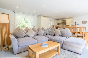 A modern living room with a grey sectional sofa, a wooden coffee table, and a view of a kitchen and dining area through a large window.