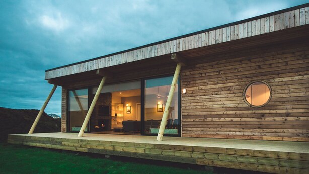A modern house with a wooden exterior, large glass windows, and a visible fireplace inside.