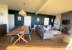 A modern living room with a wooden dining table, blue chairs, a grey sofa, and a stone fireplace.