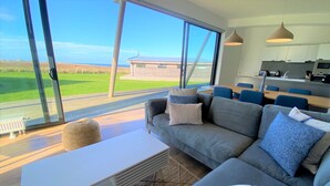A modern living room with a large window, a sofa, a coffee table, and a kitchen area visible through the glass.