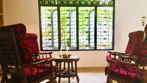 Une pièce comprenant deux chaises en bois à l’assise recouverte d’un tissu à motifs rouges, une petite table ronde et une fenêtre ornée de barreaux en métal décoratifs.