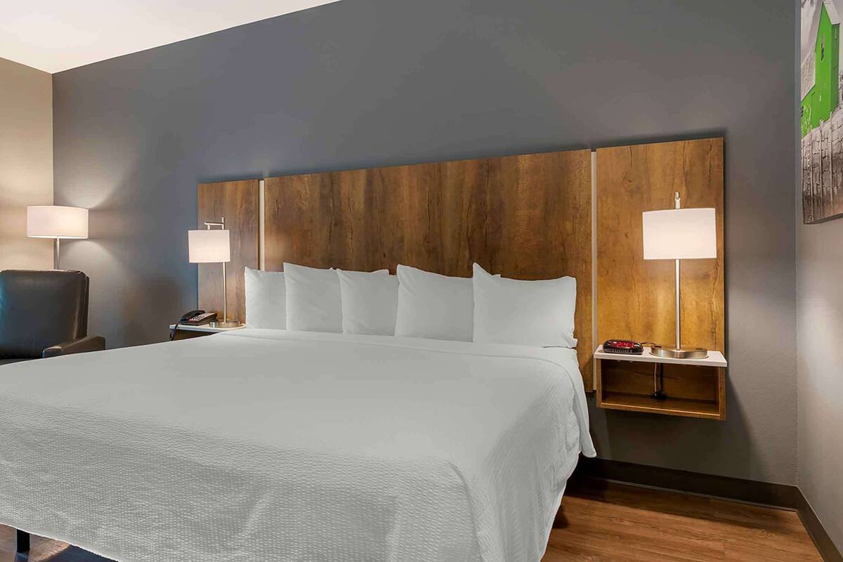 A hotel room with a large bed, a nightstand with a lamp, a chair, and a wall-mounted shelf with a lamp.