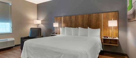 Habitación de hotel con una cama grande, una mesita de noche con lámpara, una silla y una estantería de pared con lámpara.
