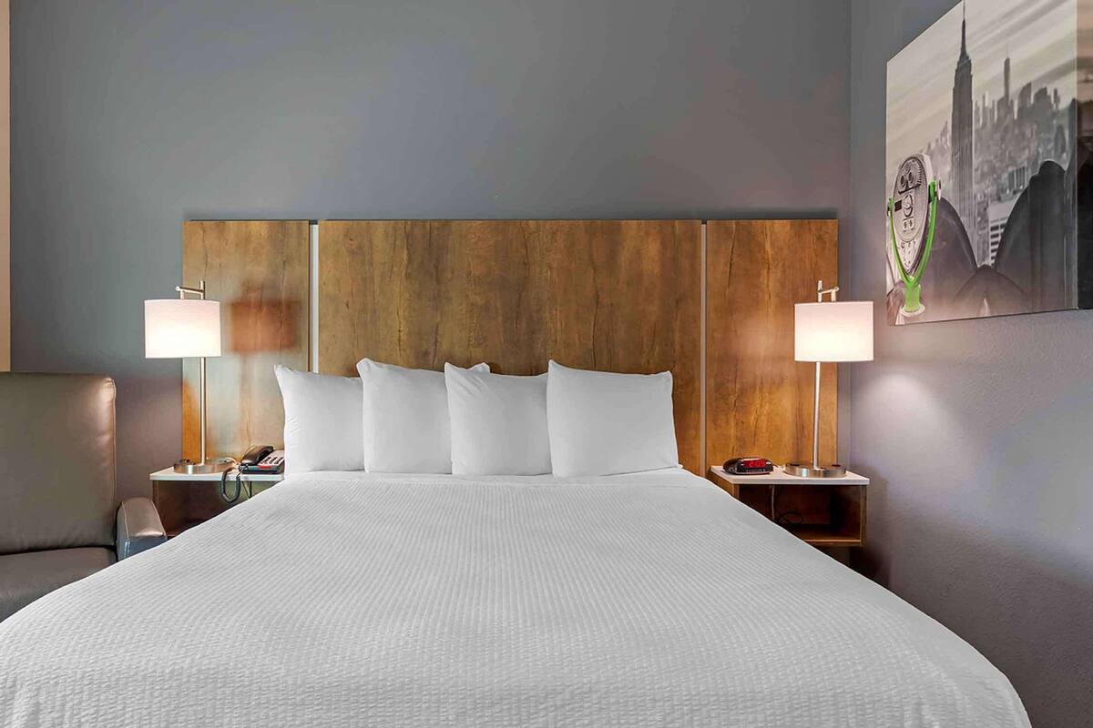 A hotel room with a large bed, two bedside tables with lamps, a chair, and a cityscape painting on the wall.