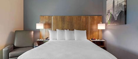 A hotel room with a large bed, two bedside tables with lamps, a chair, and a cityscape painting on the wall.