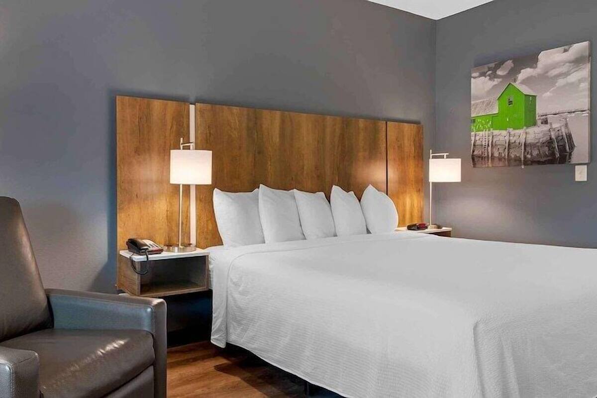 A hotel room with a large bed, a wooden headboard, a grey armchair, bedside lamps, and a painting on the wall.