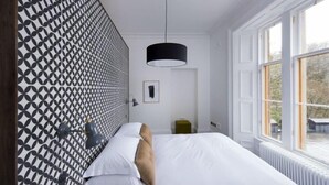 Une chambre avec une tête de lit à motifs, un lit recouvert de draps blancs et agrémenté d’un oreiller marron, une suspension noire et une fenêtre donnant sur un paysage verdoyant.