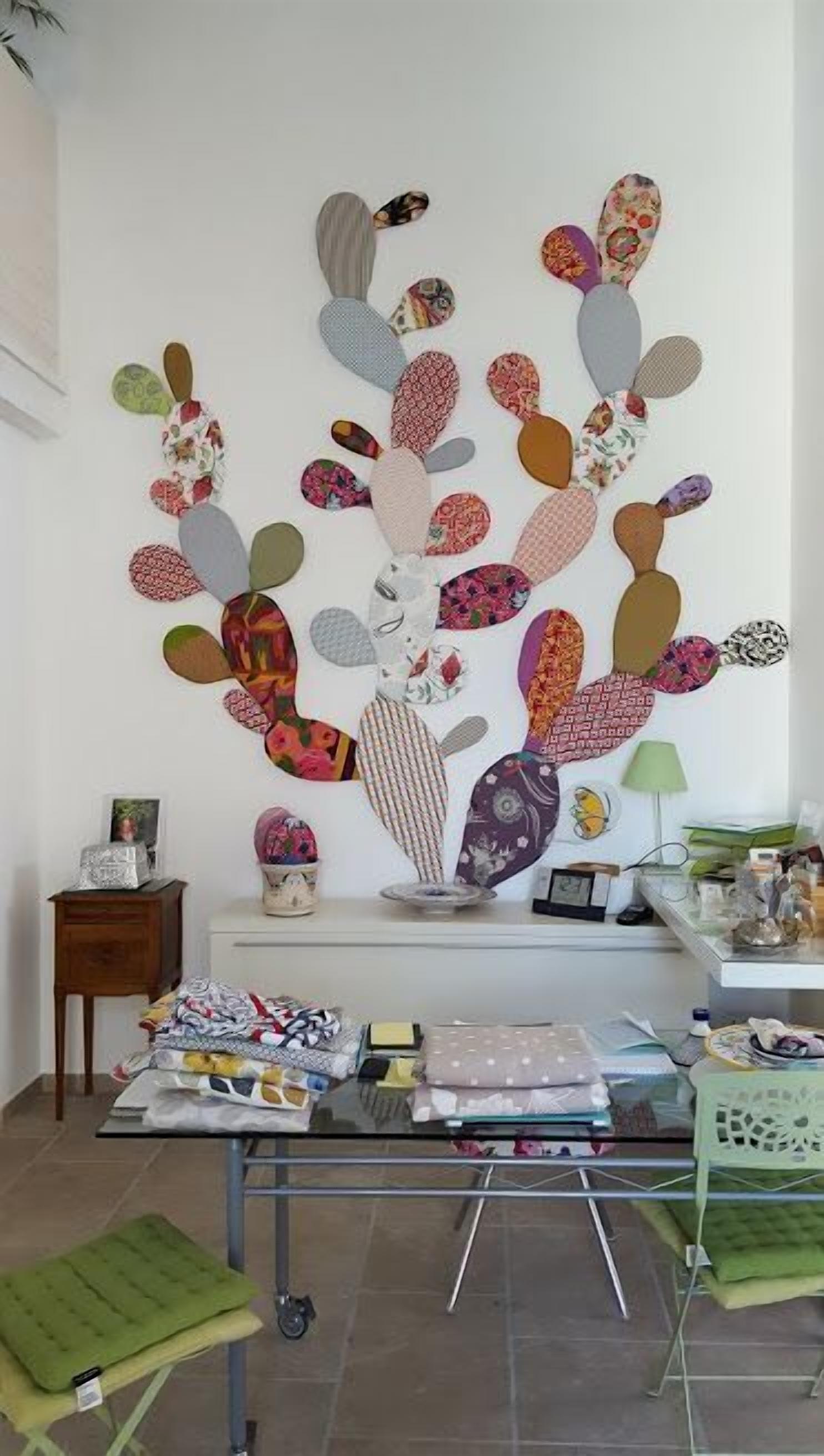 Um quarto com uma parede decorada com diversos objetos revestidos em tecido, uma pequena mesa com roupas dobradas e uma cadeira verde.