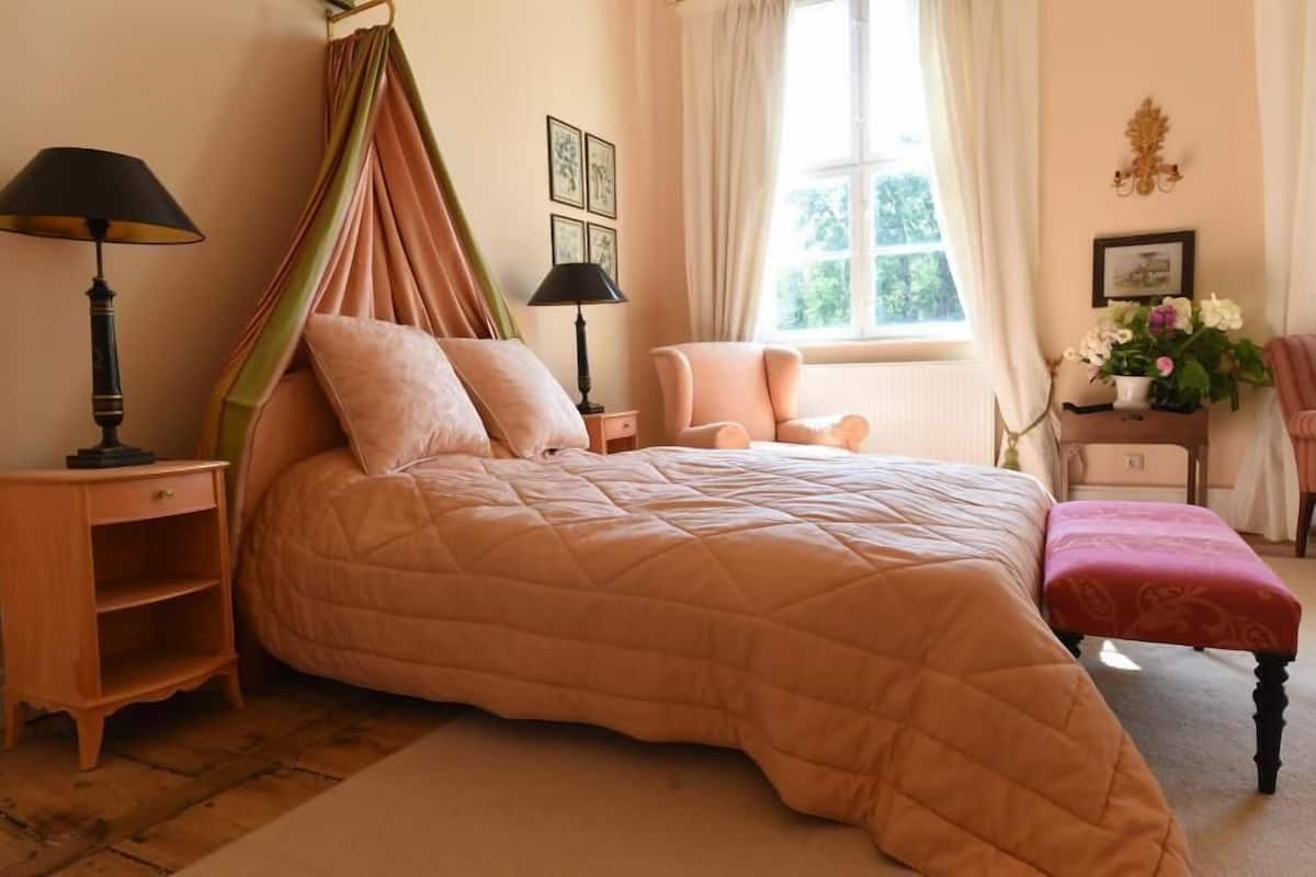 A bedroom with a large bed, two bedside tables, a window with curtains, a chair, and a small table with a vase of flowers.