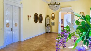 Un corridoio con pavimento decorato, porte bianche, luci a parete, quadri incorniciati e un lampadario.
