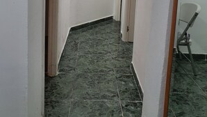 A narrow hallway with white walls and dark green marble tiles. There are wooden doors on the left and a white chair on the right.
