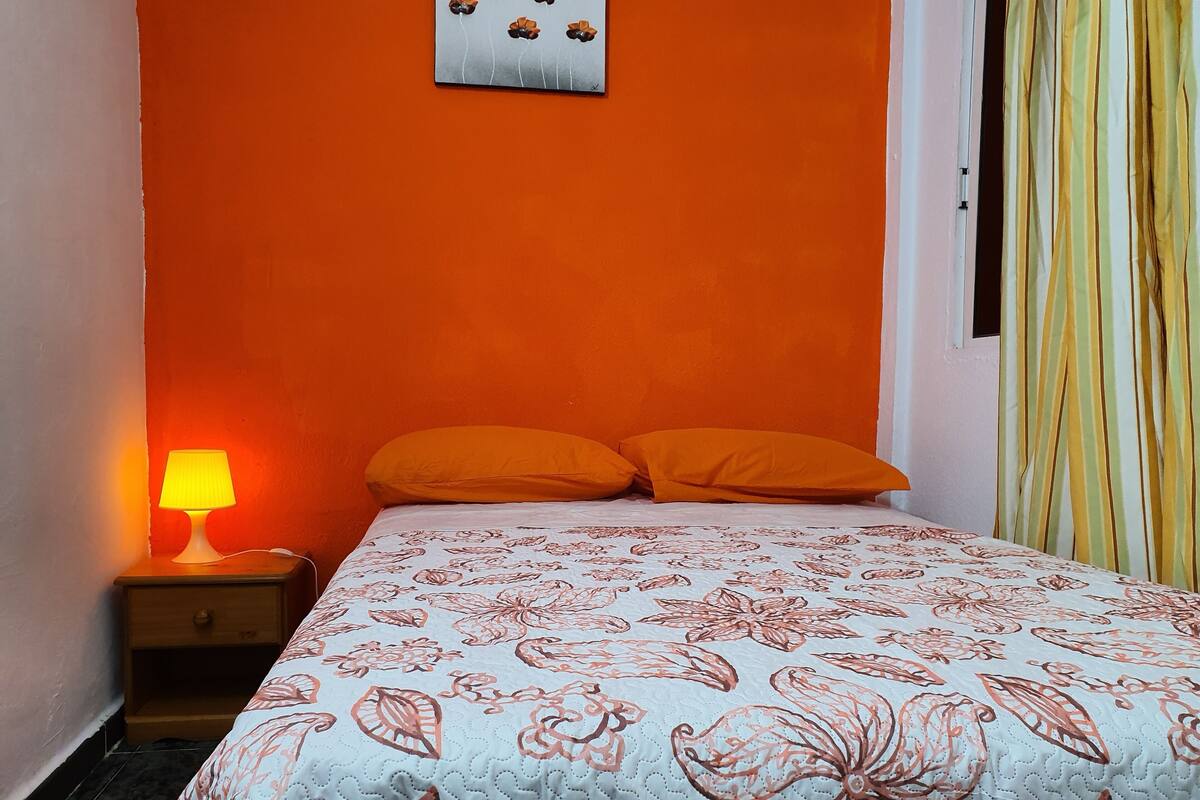 A room with an orange wall, a bed with a floral-patterned bedspread, two orange pillows, a nightstand with a lamp, and a window with striped curtains.