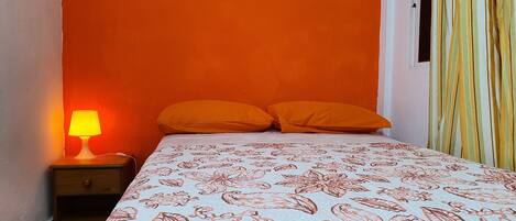 Quarto com parede laranja, cama com colcha estampada, duas almofadas laranjas, mesa de cabeceira com luminária e janela com cortinas listradas.