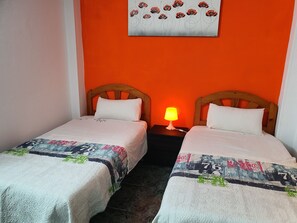 A room with two beds, each with a white pillow and a patterned blanket. There is a nightstand with a lamp between the beds.