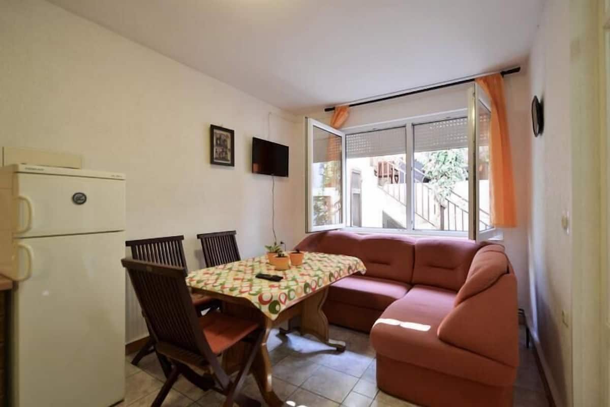 Ein gemütlicher Wohnraum mit Essbereich, einer Couch, einem kleinen Tisch mit gemustertem Tischläufer und einem Fernseher an der Wand.