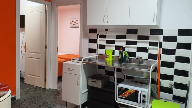 A compact kitchen with white cabinets, a microwave, and a sink. There's a small dining area with a bed in the background. - Low Cost Room Plaza de Hierro II (San Bartolomé de Tirajana)