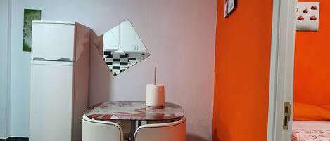 Une pièce de taille modeste, avec une petite table, deux chaises, un réfrigérateur blanc, un miroir et un mur orange.