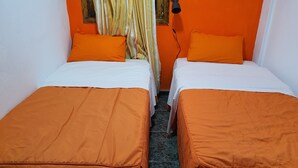 A room with two beds, each with an orange bedspread and white pillows, a window with striped curtains, and a wall-mounted lamp. - Low Cost Room Plaza de Hierro II (San Bartolomé de Tirajana)