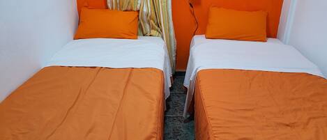 Une chambre avec deux lits, chacun recouvert d’une couverture orange et agrémenté d’oreillers blancs, une fenêtre avec des rideaux à rayures et une lampe fixée au mur.