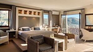 A spacious hotel room with a large bed, a sitting area with wicker furniture, a small desk, and a balcony with a scenic view. - De Tafelberg Guesthouse (Cape Town)