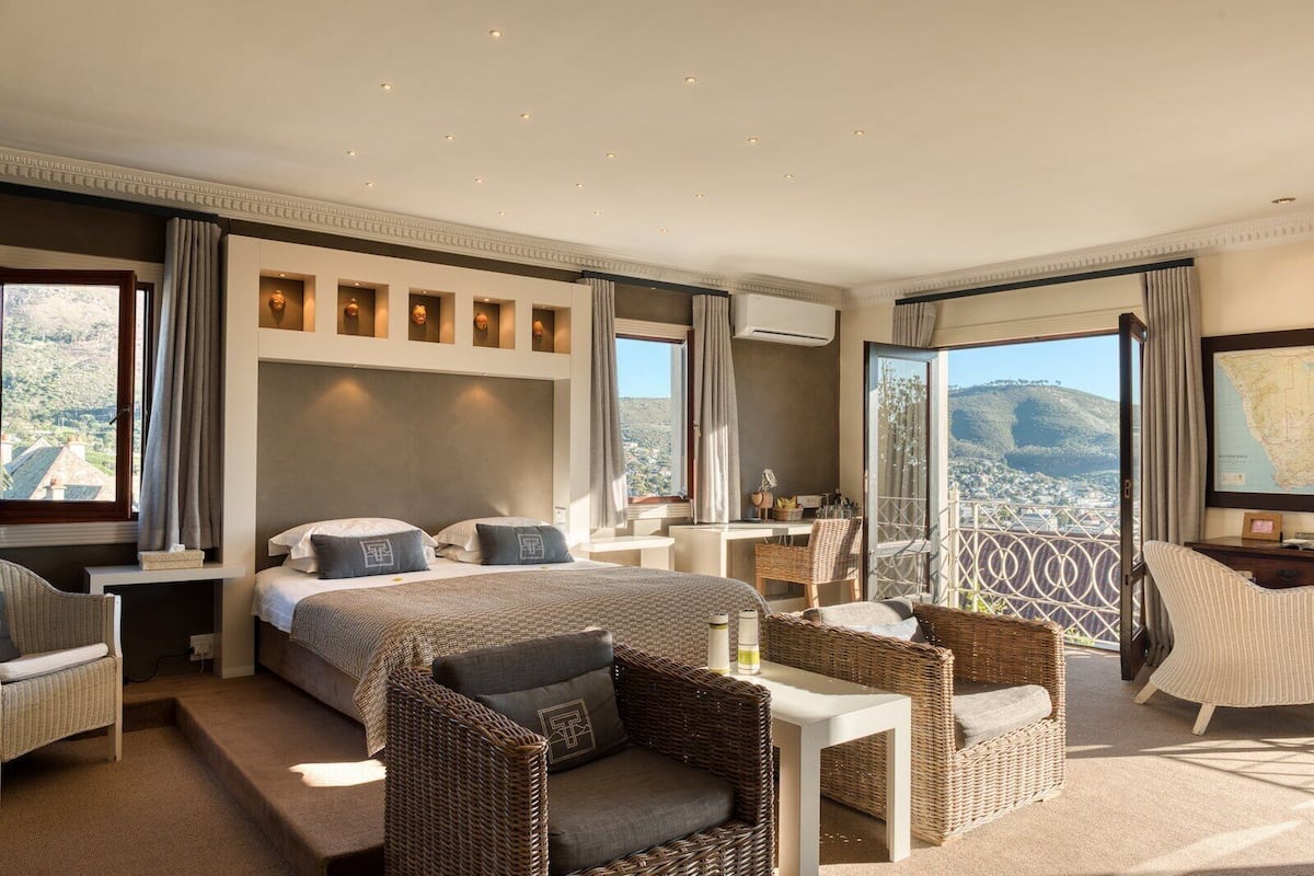 A spacious hotel room with a large bed, a sitting area with wicker furniture, a small desk, and a balcony with a scenic view.