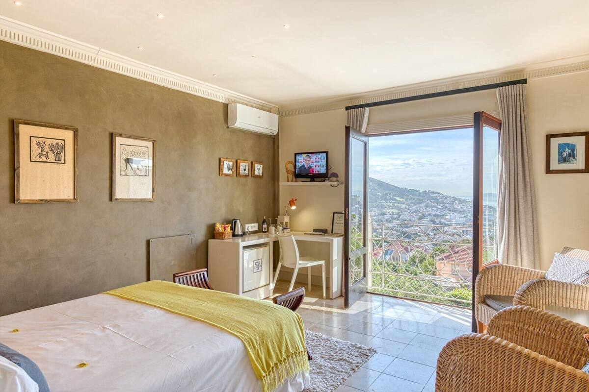 A bedroom with a bed, a desk, a chair, and a view of a cityscape through a balcony.